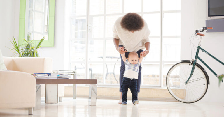 Mother helping young boy walk in a modern living room.